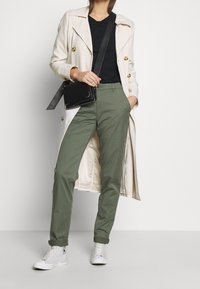 Beige dubbelgeknoopt trenchcoat over een zwart T-shirt, gecombineerd met olijfgroene broek en witte sneakers, met een zwarte crossbodytas in de hand.