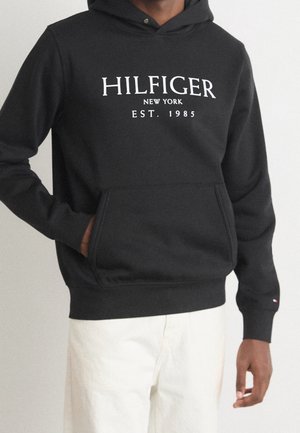 Person trägt einen schwarzen Hoodie mit weißem "HILFIGER NEW YORK EST. 1985" Schriftzug und weiße Hose, Hand in der Kapuzenjackentasche.