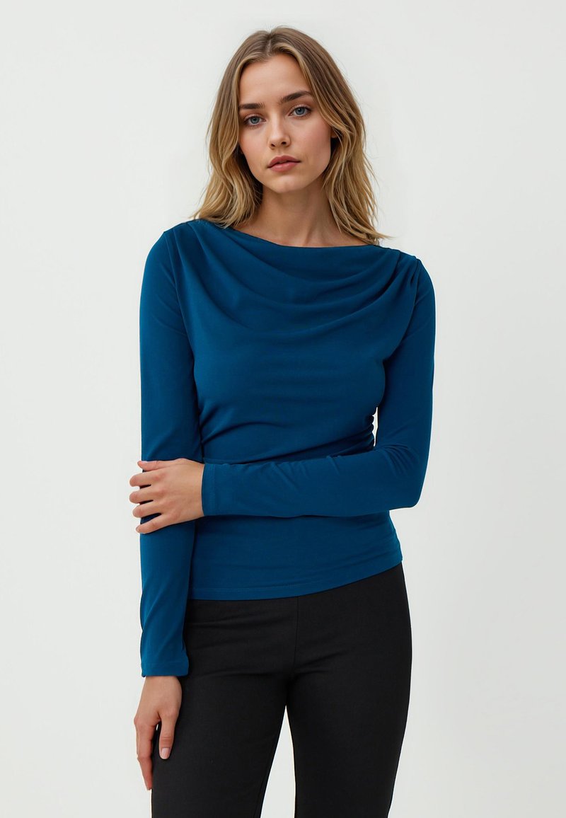Maglietta a maniche lunghe, drappeggiata, di un profondo blu teal, realizzata in morbido tessuto. Presenta un collo a cowl e una silhouette aderente, abbinata a pantaloni neri.
