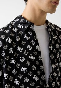 Chemise noire avec motifs graphiques blancs, comprenant des logos et des formes géométriques ; tissu léger, col boutonné, coupe décontractée.