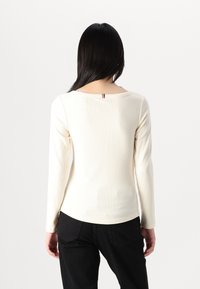Cremfarbener, gerippter Langarmshirt mit rundem Halsausschnitt und einem kleinen farbigen Akzent auf der Rückseite, kombiniert mit schwarzen Hosen.