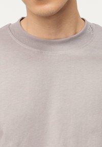 Κοντινό πλάνο ενός ατόμου που φοράει ένα ανοιχτό γκρι μπλουζάκι με στρογγυλή λαιμόκοψη και ένα μικρό κεντητό λογότυπο κοντά στον γιακά.