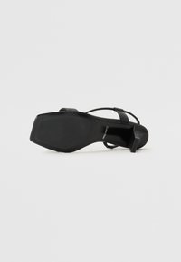 Sandalie negre cu toc înalt, cu un talpă netedă și texturată și o formă unghiulară. Prezintă bretele înguste și un design minimalist.