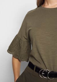 T-shirt vert olive avec des manches courtes à volants brodés, associé à une ceinture noire ornée de boucles en argent superposées.