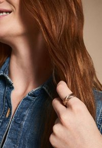 Ring mit einem Chevron-Muster aus roségold, vor einem Hintergrund aus einem Jeanshemd mit blauen Knöpfen und langen, glatten roten Haaren.