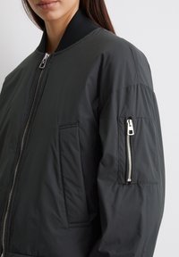 Schwarze Bomberjacke mit einem gerippten Kragen, Reißverschluss vorne und einer Seitentasche. Sie hat eine glatte Stofftextur und einen leicht oversized Schnitt.