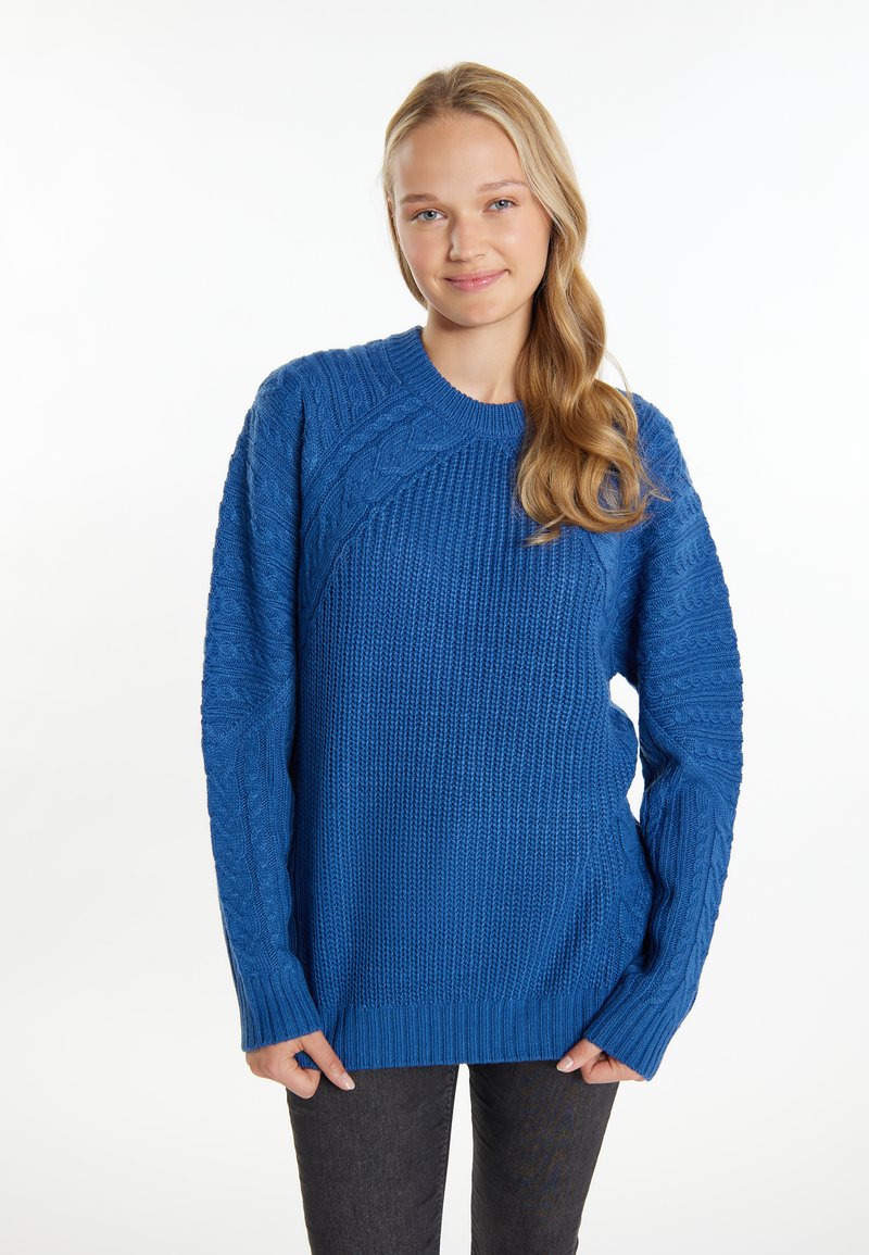 Maglione azzurro a maglia con motivi testurizzati, colletto a girocollo e maniche lunghe con polsini a coste. Indossato sopra jeans scuri.