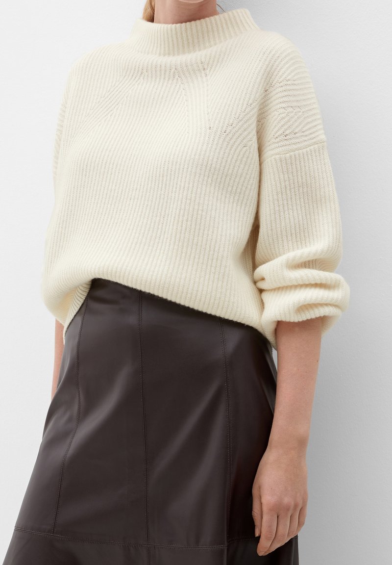 Suéter de punto acanalado en color crema con cuello alto y mangas enrolladas, combinado con una falda de cuero marrón oscuro con detalles de costura.
