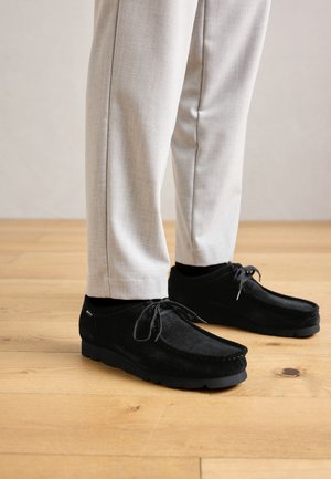 Jambes portant un pantalon beigne clair et des chaussures en daim noires à lacets, debout sur un sol en bois.