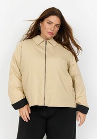 Bežna zip-up jakna s ovratnikom, črnimi manšetami in sproščenim krojem. Gladek material z lahkotnim in sproščenim dizajnom.