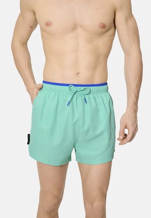 Hombre sin camiseta con pantalones de baño de color turquesa claro con cintura y cordón azules, de pie con una mano en el bolsillo frente a un fondo blanco.