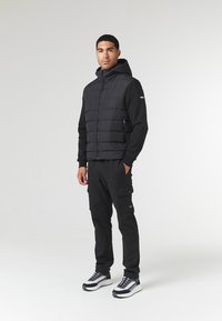 Sort pufferjakke med hætte og lynlås, båret over en sort langærmet skjorte og cargo bukser. Har kontrasterende grå sneakers.