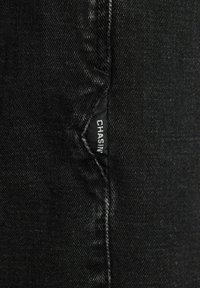 Tela de mezclilla negra con una textura suave y ligero brillo, que presenta una etiqueta de marca visible "CHASIN" cosida en la costura.