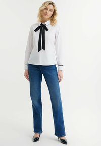 Blusa blanca de manga larga con una cinta negra atada en el cuello, combinada con jeans rectos azules y tacones negros de punta.