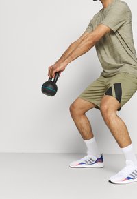 Kettlebell w kolorze czarnym z niebieskimi akcentami trzymany przez osobę w jasnej oliwkowej koszulce sportowej i szortach z czarnymi detalami, w białym obuwiu.