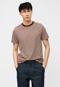 Νεαρός άνδρας που φοράει κοντομάνικη μπλούζα με ρίγες μπορντό και μπεζ, ελαφρώς μπουκαρισμένη σε σκούρο μπλε τζιν παντελόνι, με τα χέρια στις τσέπες.