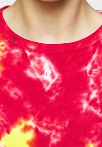 Röd tie-dye t-shirt med gula och vita mönster, med ribbad krage och v-formad detalj vid halsringningen. Mjuk bomullstyg.