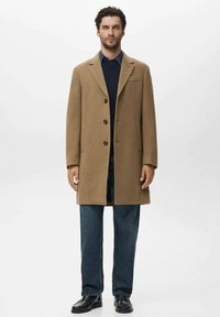 Abrigo de lana marrón con cuello con muescas, cierre de botones y dos bolsillos laterales. Combinado con un suéter azul marino y jeans de un azul oscuro.
