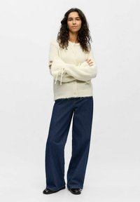Cárdigan de punto color marfil con un patrón texturizado, cierre de botones y lazos de cinta en los puños, combinado con jeans de mezclilla oscura de pierna ancha.