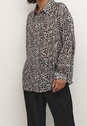 Person trägt ein langärmliges, geknöpftes Hemd mit Leopardenmuster, kombiniert mit lockeren schwarzen Hosen, und steht vor einem schlichten hellen Hintergrund.