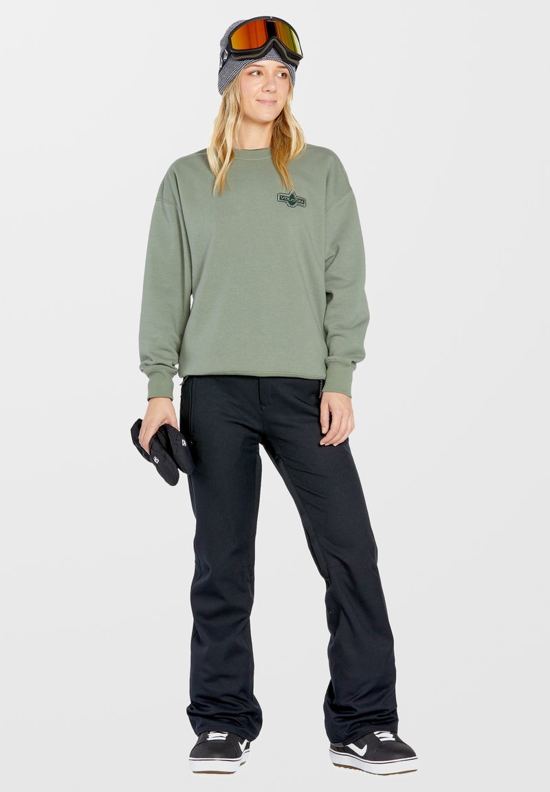 Groene sweatshirt met een logo, zwarte sneeuwbroeken en zwarte handschoenen. Het model draagt een grijze beanie en spiegelende skibril met oranje accenten.