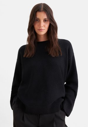 Femme aux longs cheveux bruns portant un pull noir et un pantalon noir, debout les mains dans les poches sur un fond blanc uni.