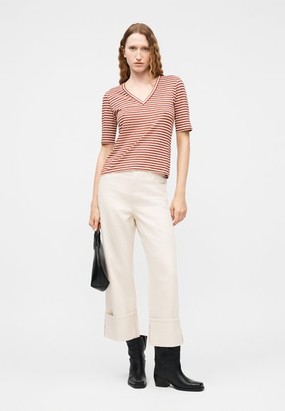 Γυναίκα με κοντομάνικο τοπ V-neck με ρίγες σε σκουριά και λευκό, φαρδιά κρεμ παντελόνια με μανσέτα, μαύρα μποτάκια αστραγάλου, κρατώντας μικρή μαύρη τσάντα.