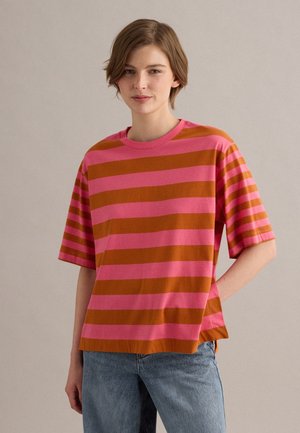Giovane persona che indossa una maglietta larga a righe rosa e arancione e jeans blu, in piedi con una mano in tasca davanti a uno sfondo uniforme.