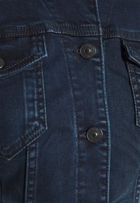 Tmavě modrá džínová bunda s dvěma náprsními kapsami, černými kovovými knoflíky a detailem šití. Hladká textura s přiléhavým střiheml.