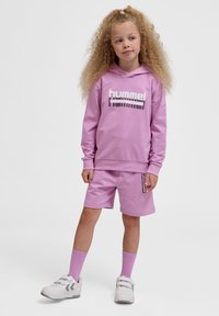 Różowa bluza z kapturem z logo "hummel", dopasowane szorty i jasnoróżowe skarpetki. Charakteryzuje się luźnym krojem i miękkim materiałem. Białe sneakersy z akcentami.