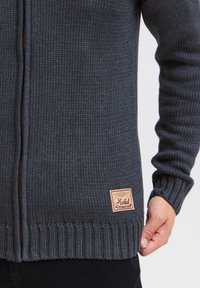 Mörkgrå stickad tröja med dragkedja, ribbade ärmslut och nederkant, samt en cirkulär läderlogotyp på nedre högra sidan.