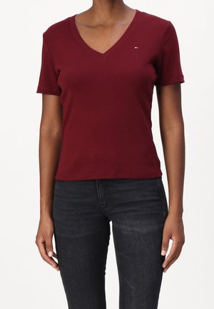 Osoba ubrana w dopasowany bordowy T-shirt z krótkim rękawem i dekoltem w serek oraz ciemnoszare dżinsy, stojąca z rozluźnionymi ramionami wzdłuż ciała.