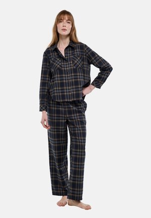 Femme portant une chemise à carreaux bleu marine et marron boutonnée et un pantalon large assorti, debout pieds nus avec une main sur la hanche.