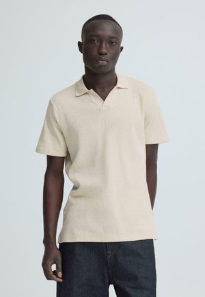 Jeune homme debout portant un polo à manches courtes beige clair texturé et un jean bleu foncé, sur un fond clair uni.