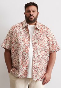 Chemise à manches courtes en tissu léger, fond crème avec motif floral rouge. Design à boutons, coupe décontractée, portée par-dessus un T-shirt blanc.