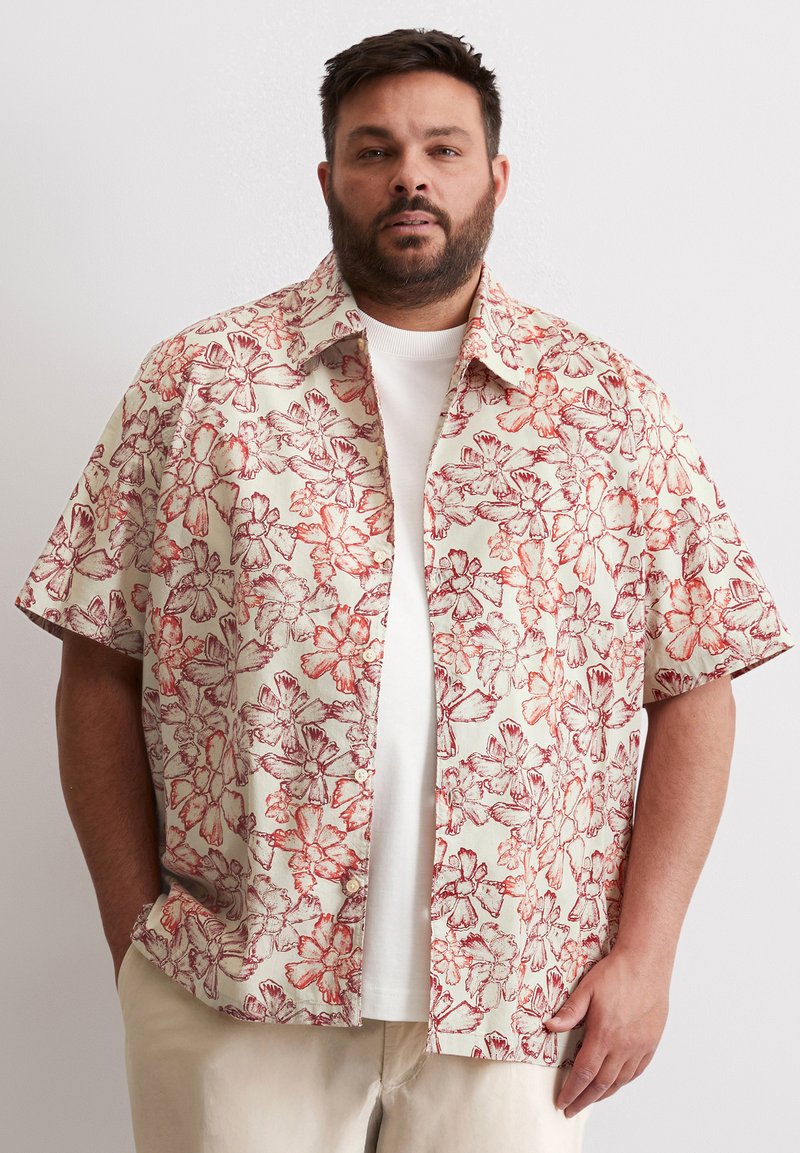 Chemise à manches courtes en tissu léger, fond crème avec motif floral rouge. Design à boutons, coupe décontractée, portée par-dessus un T-shirt blanc.