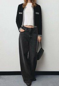 Mujer vistiendo una blusa corta blanca, chaqueta abierta negra texturizada con trim plateado, jeans negros de pierna ancha, zapatos negros y sosteniendo un pequeño bolso negro.