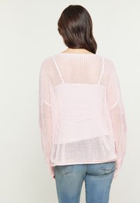Maglione in maglia oversized di un rosa chiaro, con una trama allentata. Presenta spalle scese e orlo ampio. Indossato sopra un paio di jeans blu chiaro.