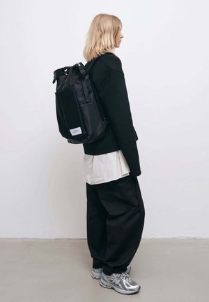 Sort rygsæk med rullegardinsdesign, tekstureret stof, justerbare stropper og en frontlommepose i net. Båret over et sort outfit.