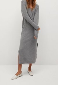 Femme portant une longue robe tricotée grise avec des fentes sur les côtés et des chaussures plates blanches, se tenant devant un fond clair.