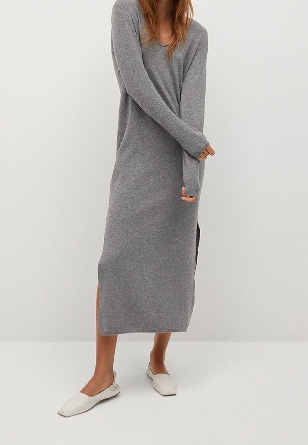 Femme portant une longue robe tricotée grise avec des fentes sur les côtés et des chaussures plates blanches, se tenant devant un fond clair.
