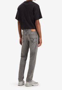 Vaqueros de mezclilla gris con corte ajustado, que presentan un parche de cuero en la parte posterior, dos bolsillos traseros y un ligero desgaste. Combinados con una camiseta negra de manga corta.