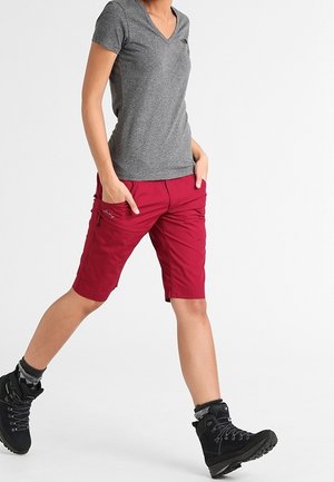 Szara koszulka z dekoltem w serek w połączeniu z czerwonymi szortami do kolan z zamykanymi na zamek kieszeniami oraz czarne buty trekkingowe z teksturowanymi detalami.