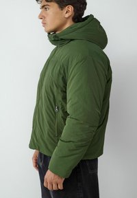 Giacca a vento verde con cappuccio, caratterizzata da una zip frontale e una tasca laterale con zip. Realizzata in materiale leggero e testurizzato.