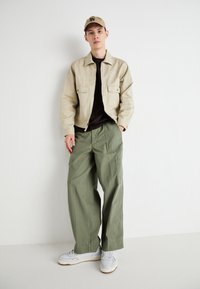 Light beige zip-up jacket with two front pockets, paired with olive green wide-leg pants and white sneakers. Set against a plain background.