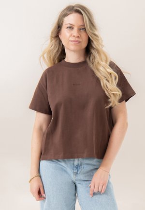 Kvinde med langt blond hår iført en brun kortærmet skjorte og lyseblå jeans, stående mod en ensfarvet lys baggrund.