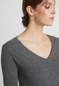 Pilkas ilgas rankovėmis V formos iškirptės marškinėliai iš minkštos, tekstūruotos medžiagos, derinami su paprasta auksine auskaru žiedo formos. Marškiniai turi laisvą fasoną aplink iškirptę.