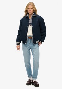 Chaqueta varsity de lana azul marino con puños a rayas, llevada sobre un suéter crema y una camisa con cuello, combinada con jeans azul claro y mocasines burdeos.