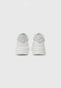 Adidași cu platformă albă din piele netedă, având vârful rotund și un detaliu proeminent în spate cu un logo și accente decorative cu capse.