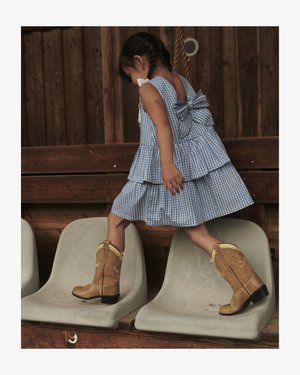 Kék mintás ruhát viselő gyermek és világosbarna cowboy csizmák, amint üres műanyag székek között lépked egy fa pajtában.